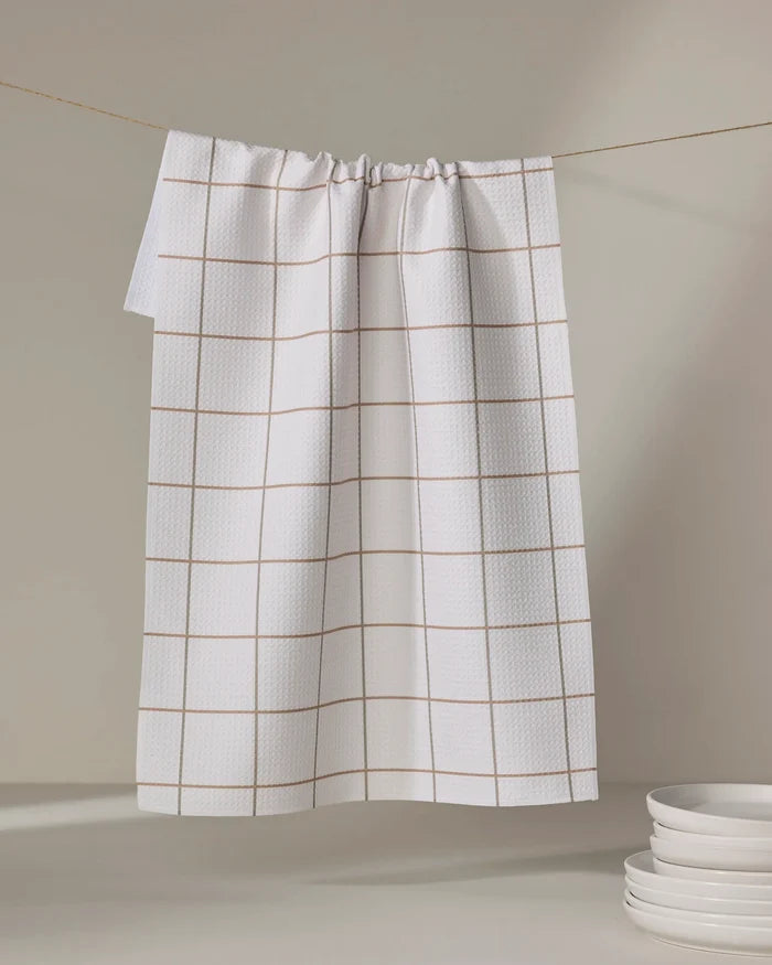 The Ivory Grid tea towel by Kelsi Fullmer, featuring a light brown grid on white, hangs on a line with stacked white plates in the corner against a neutral background.