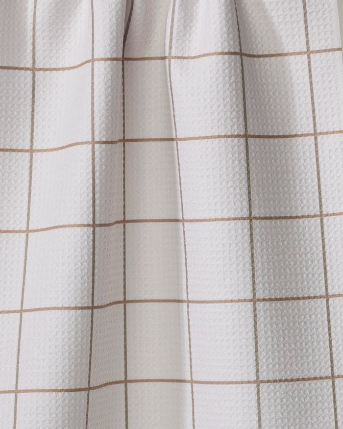 The Kelsi Fullmer Ivory Grid kitchen towel features a light-colored fabric with thin, evenly spaced brown lines in a grid pattern, draped to create soft folds and subtle shadows.