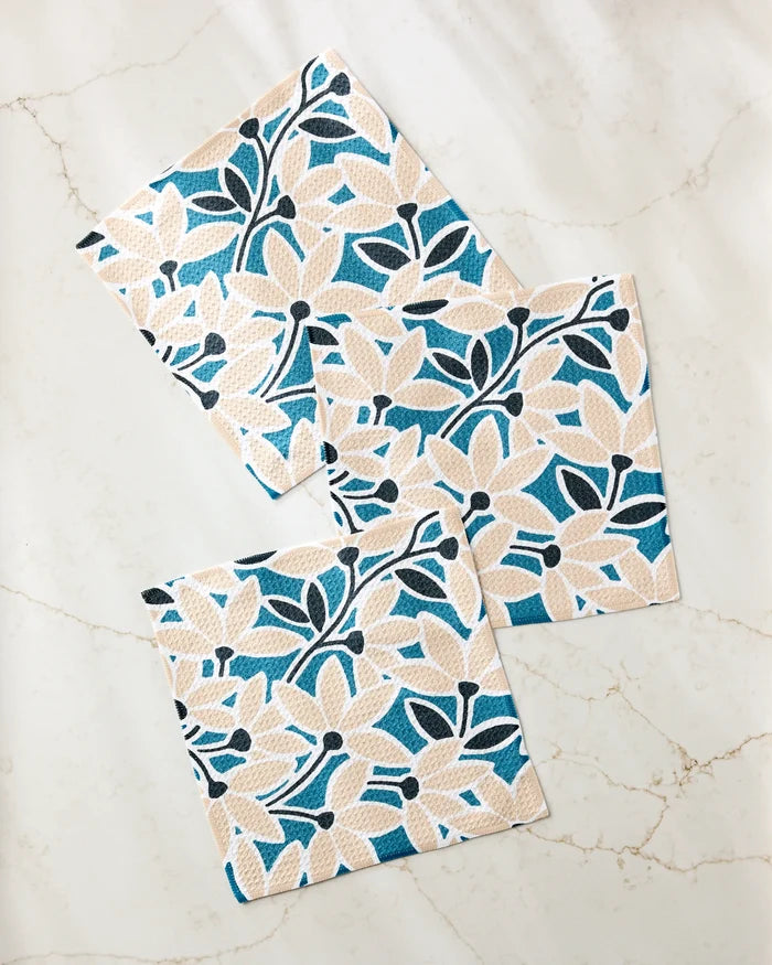 Three Janine dishcloths by Amy MacCready—beige with blue and black floral patterns—are arranged on a white marble surface with subtle grey veining, slightly overlapping and placed at varying angles.
