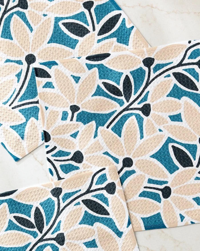 The Janine dishcloth set by Amy MacCready features three overlapping absorbent cloths with beige flowers, black stems, and a teal-blue background, displayed on light marble—perfect for brightening up any kitchen.