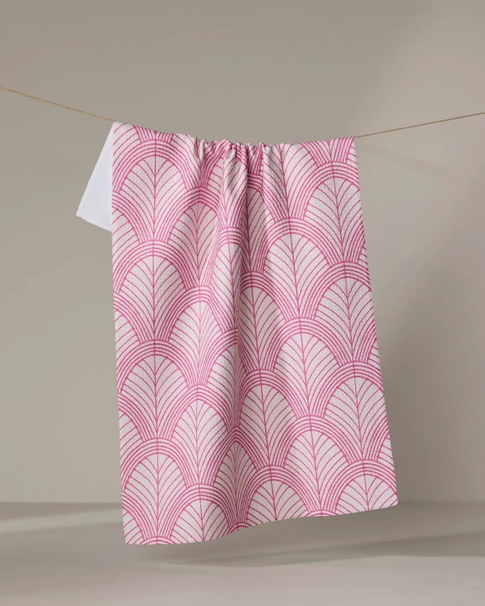 The Jaskin dish towel by Amy MacCready, with a pink geometric fan design on white, hangs on a clothesline against a light gray background.