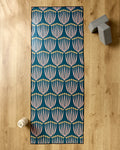 The Josie yoga mat by Amy MacCready, featuring pink and yellow floral patterns on teal, is displayed on a wooden floor with a non-slip yoga towel, two gray yoga blocks, and a white water bottle.