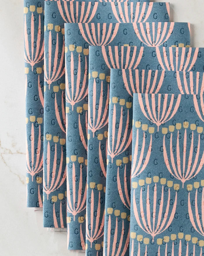 Five Josie napkins by Amy MacCready, stacked in a fan arrangement, showcase pink and yellow stylized menorahs on blue cloth with subtle “G” monograms—an eco-friendly alternative to paper towels.