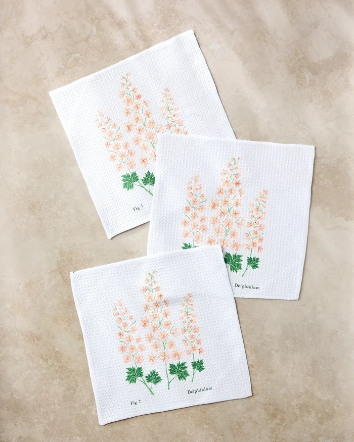 Three July Delphinium napkins by Linda Fox, made from recycled materials, display peach and green delphinium floral illustrations labeled Fig 1 or Delphinium on a light beige surface.