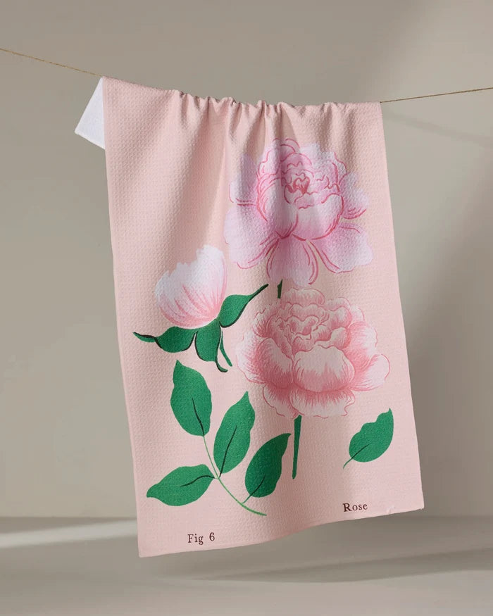 The June Roses tea towel by Linda Fox is pale pink, made from recycled materials, and features pink rose and green leaf illustrations. "Fig 6" and "Rose" are labeled at the bottom on a neutral background as it hangs on a line.