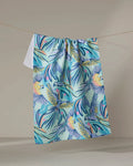 The "Jungle Birds" tea towel by Masha Volnova features a vibrant tropical design with parrots, palm leaves, and sun motifs in blue, yellow, green, and pink. Made from recycled materials, it's shown hanging on a neutral background.