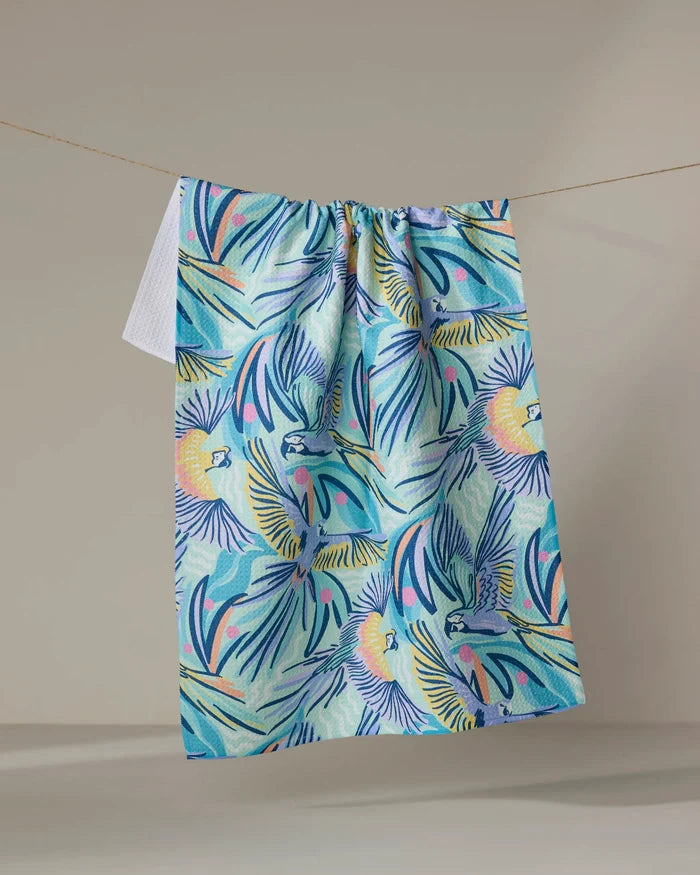 The "Jungle Birds" tea towel by Masha Volnova features a vibrant tropical design with parrots, palm leaves, and sun motifs in blue, yellow, green, and pink. Made from recycled materials, it's shown hanging on a neutral background.