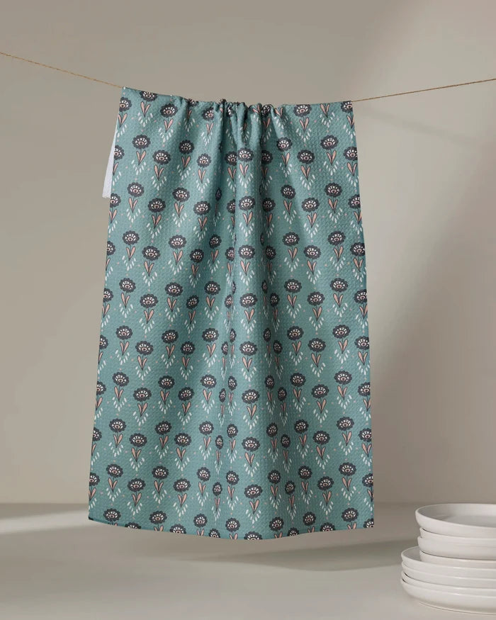 The "Jungle Petals" tea towel by Amita Nair, featuring teal floral patterns and made from recycled materials, hangs against a neutral backdrop, with a stack of white plates in the lower right corner.