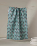 The "Jungle Petals" tea towel by Amita Nair, featuring teal floral patterns and made from recycled materials, hangs against a neutral backdrop, with a stack of white plates in the lower right corner.