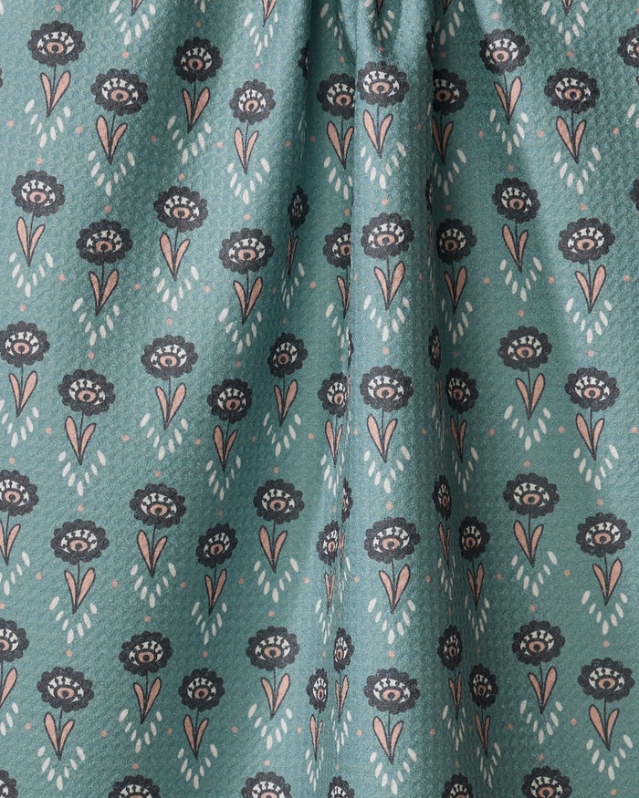 The Jungle Petals tea towel by Amita Nair features black and pink floral motifs with leaves on teal fabric, arranged in rows. Slight top gathering adds charm to this stylish and functional kitchen essential.