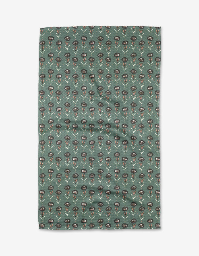 The Jungle Petals tea towel by Amita Nair features green Geoweave fabric with a pattern of small, stylized red, black, and beige flowers, shown flat with slight wrinkles.