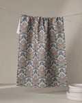 Amita Nair's "Jungle Sunrise" tea towel, made from recycled materials and ultra-absorbent, features vibrant geometric and floral patterns. It hangs by white plates stacked against a softly lit neutral background.