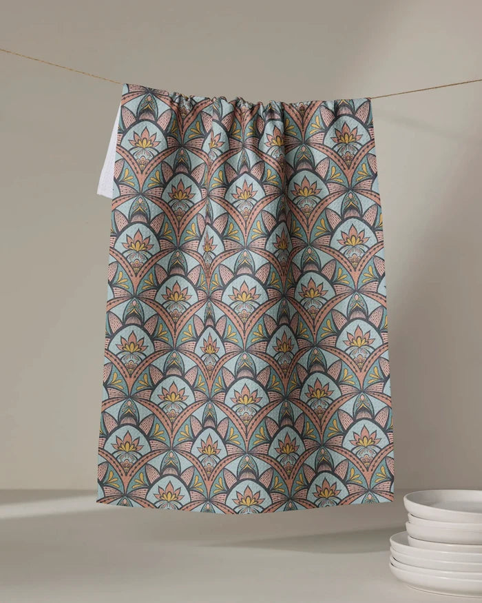 Amita Nair's "Jungle Sunrise" tea towel, made from recycled materials and ultra-absorbent, features vibrant geometric and floral patterns. It hangs by white plates stacked against a softly lit neutral background.