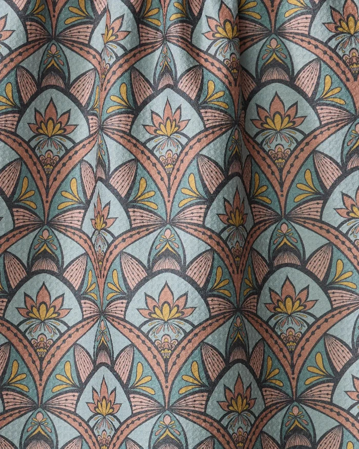A close-up of the "Jungle Sunrise" tea towel by Amita Nair, made from recycled materials. It features a repeating art deco floral pattern in yellow, peach, and black on a blue-green background with gentle folds and slight texture.
