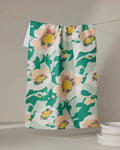 The Geometry Junie dish towel, featuring a bold floral pattern in green, yellow, peach, and white, hangs on a clothesline above a white countertop with stacked white dishes beside it.