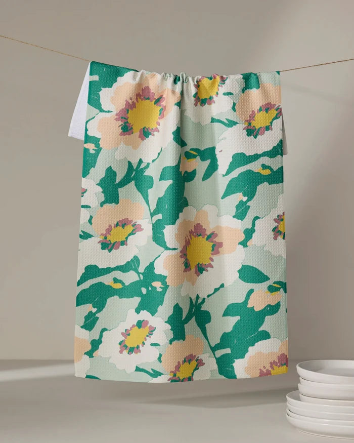 The Geometry Junie dish towel, featuring a bold floral pattern in green, yellow, peach, and white, hangs on a clothesline above a white countertop with stacked white dishes beside it.