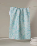 The Katie O'Shea "Just Keep Swimming" light blue quick-drying tea towel with a wavy blue line pattern hangs on a string near a stack of white plates, set against a neutral background.