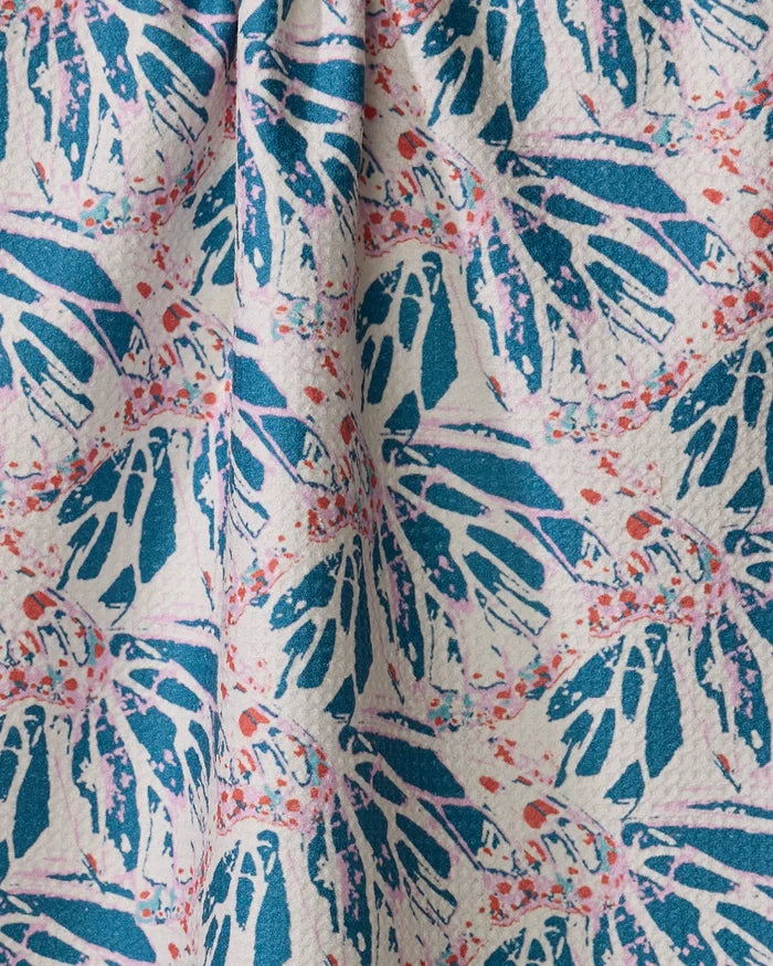 A close-up of the Rebecca Woolbright Kaleidoscope Dream kitchen towel shows a textured surface with colorful blue, pink, and coral abstract leaf patterns on a light background and a slight vertical center fold.