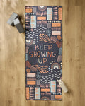 The Jessica Swift "Keep Showing Up" patterned yoga mat is on a wooden floor, accompanied by a recycled yoga towel, two gray yoga blocks, and a white water bottle.