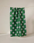 The GEOMETRY Kick Off eco-friendly kitchen towel in green features a repeating black and white soccer ball pattern and hangs on a line against a plain, light-colored background.