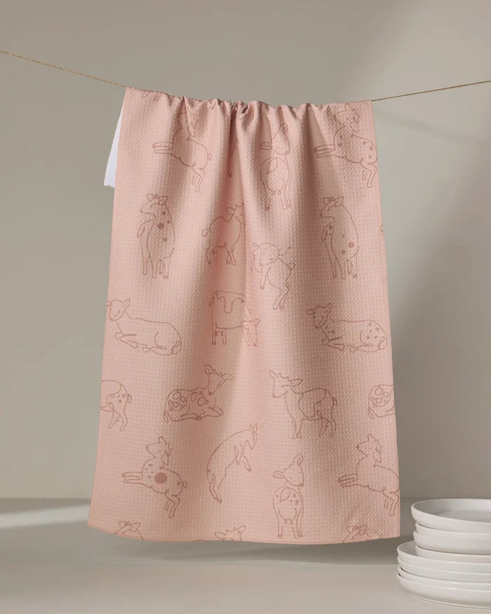 The Kidding Around tea towel by Sara Mari features pink fabric with simple cow outlines. Hanging on a string beside white plates, it adds charm and ultra-absorbent performance to everyday kitchen tasks.