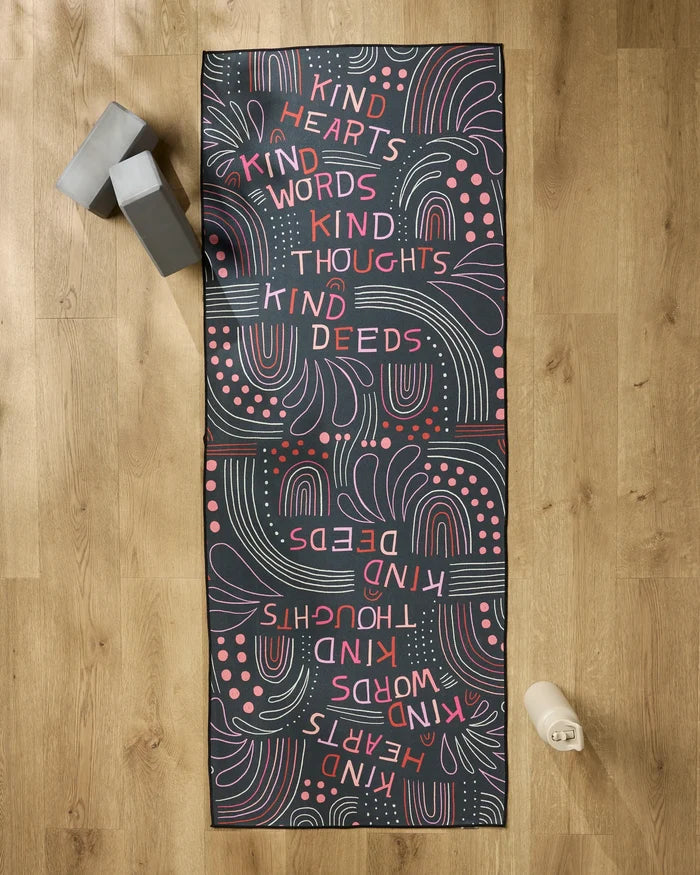 The Jessica Swift "Kind Hearts" yoga mat, red and white with the words kind hearts, kind words, kind thoughts, and kind deeds, rests on a wooden floor with gray yoga blocks, a white water bottle, and a non-slip towel beside it.