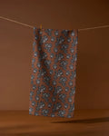 The Geometry Kindred towel, made from recycled materials, features a brown cloth with a repeating black leaf and acorn pattern. It hangs from a string with two clothespins against a brown background.