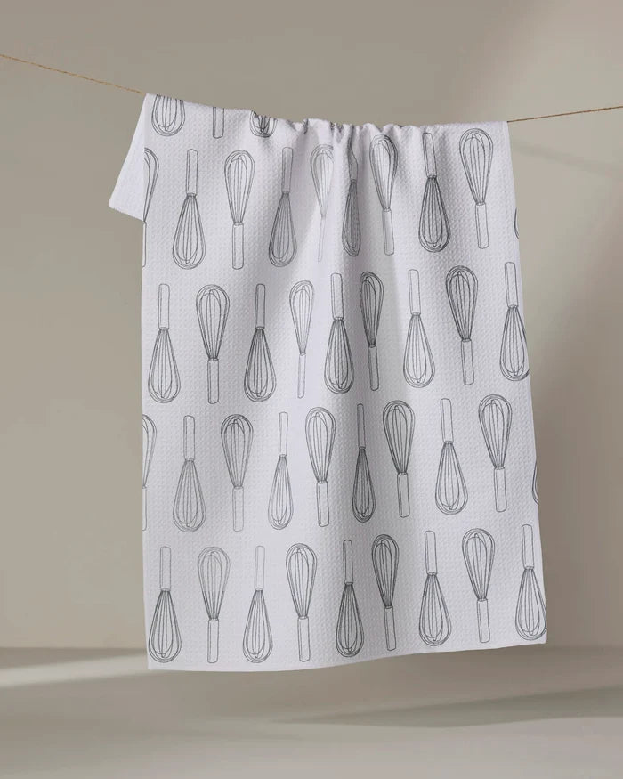 A “Kitchen Whisks” tea towel by Rosa Kichline, made from quick-drying recycled materials, hangs on a line. It features a repeating black wire whisk pattern on white and is set against a softly lit neutral background.