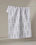 A “Kitchen Whisks” tea towel by Rosa Kichline, made from quick-drying recycled materials, hangs on a line. It features a repeating black wire whisk pattern on white and is set against a softly lit neutral background.