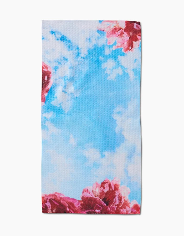 The La Floraison bar towel by Stefanie Bales showcases a bright blue sky, white clouds, and vivid pink flowers along the edges, crafted from ultra-absorbent Geoweave fabric for both style and functionality.