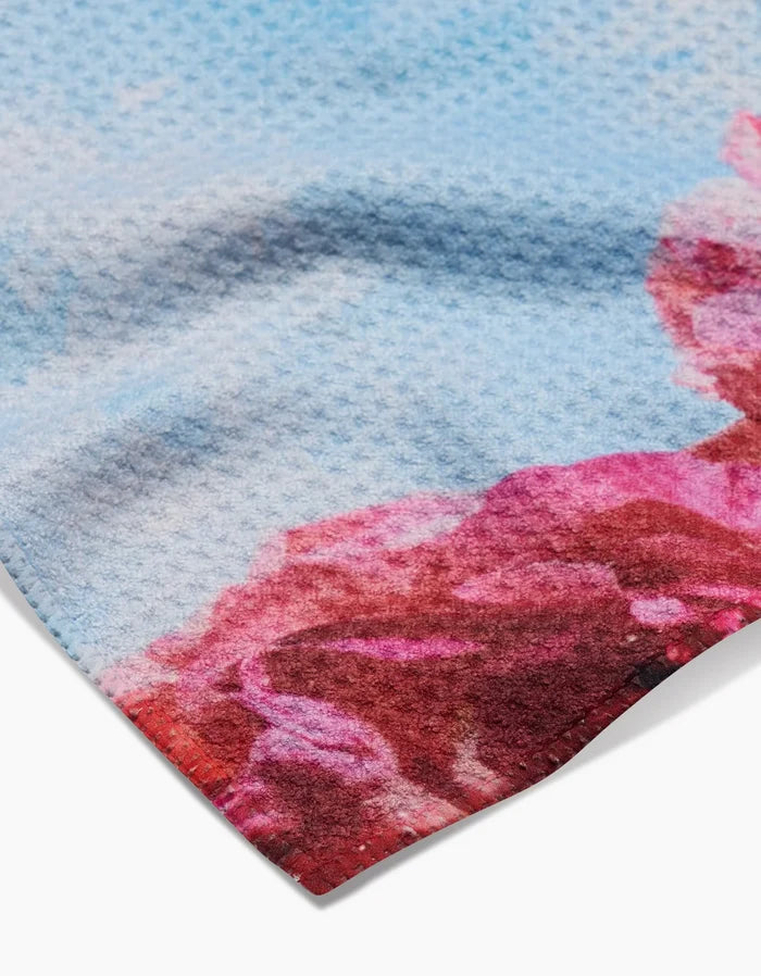A close-up of the Stefanie Bales La Floraison towel, featuring ultra-absorbent Geoweave fabric with a waffle texture and red and pink floral-inspired patterns on a light blue background.