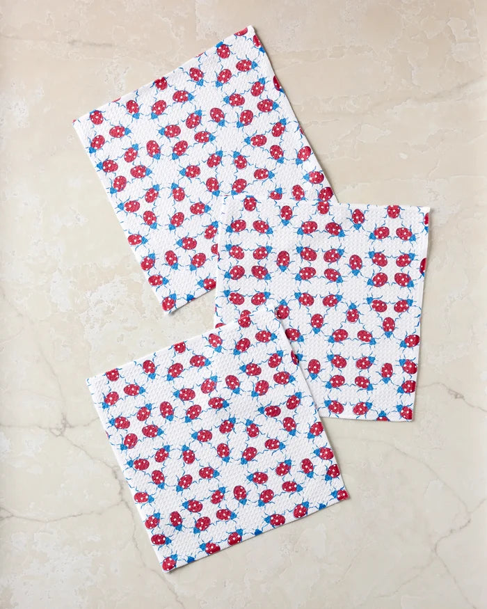 Three 'Ladybug Dance' cloth napkins by Second Nature Studio, featuring a red and blue floral pattern, are neatly folded and overlap slightly on a light beige marble surface.
