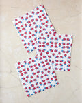 Three 'Ladybug Dance' cloth napkins by Second Nature Studio, featuring a red and blue floral pattern, are neatly folded and overlap slightly on a light beige marble surface.