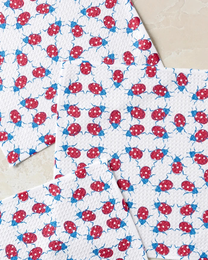 Four white "Ladybug Dance" kitchen towels by Second Nature Studio, featuring a red and blue ladybug pattern, are spread on a light stone surface with overlapping edges.