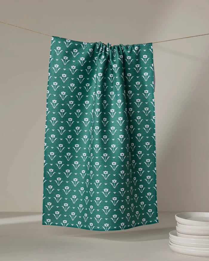 The Buy Guide's Ladylake - Evergreen recycled kitchen towel, featuring a white floral pattern, hangs on a string near stacked white plates against a neutral backdrop.