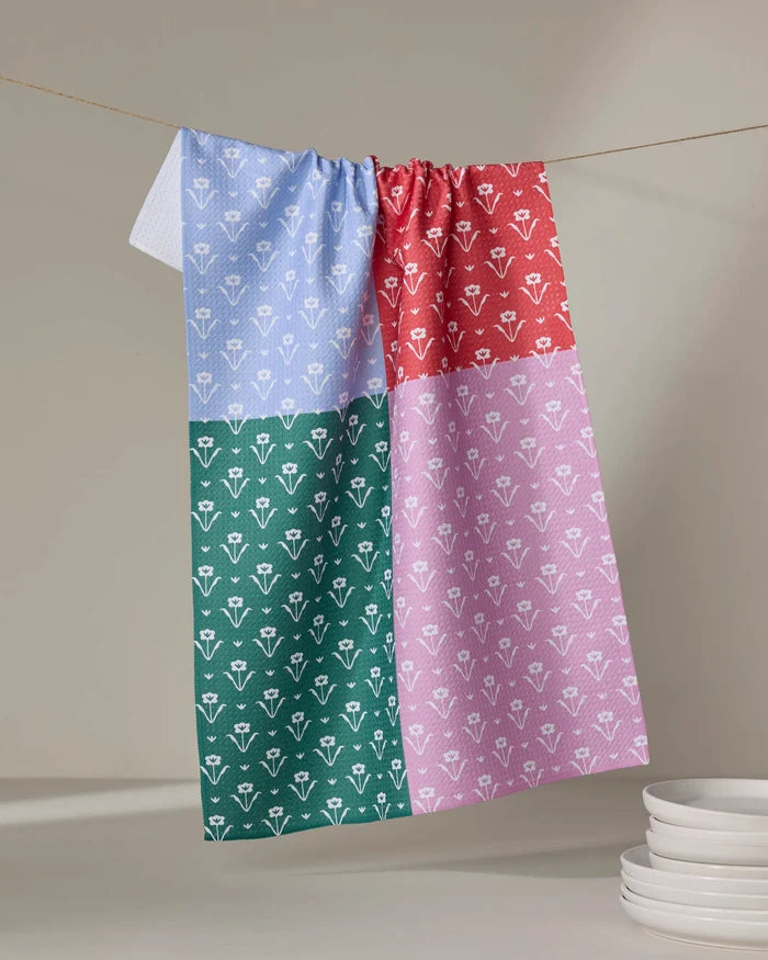 The Buy Guide’s Ladylake – Patchwork tea towel, featuring a four-color block design with a white floral pattern, hangs on a line above stacked white plates.