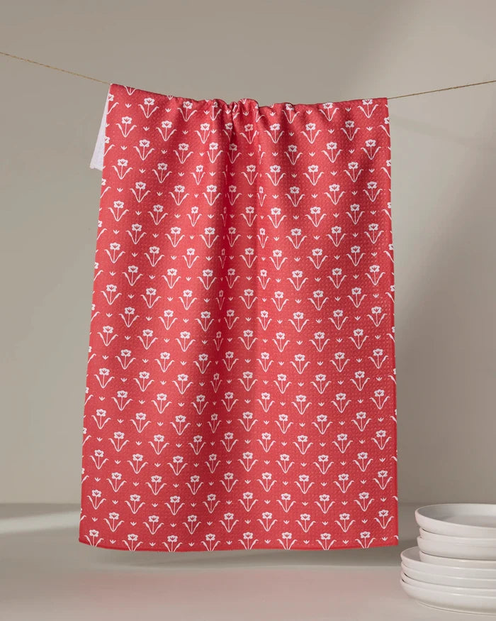A Ladylake - Red kitchen tea towel by The Buy Guide, featuring a white floral pattern, hangs on a string. Nearby, a small stack of white plates rests on a light surface.