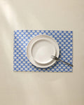 A white plate and small bowl stacked with a fork on the edge sit on a cream tablecloth over stylish Geometry Lagoon Placemats in blue and white patterns.