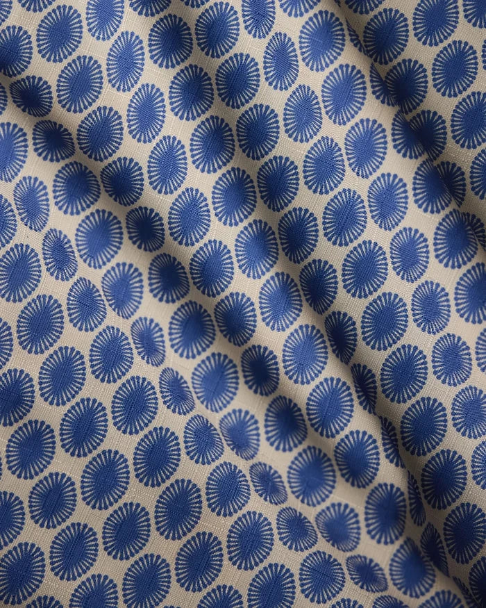 A close-up of Lagoon by Geometry shows beige fabric with a repeating blue sunburst pattern, creating a geometric, textured look. The lightweight tablecloth surface is slightly wrinkled.