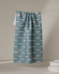The Lake Fish tea towel by Coit Creative, made from recycled materials, features teal fabric with swimming whales and pink fish. Ultra-absorbent, it's pictured hanging on a line near stacked white plates on a light surface.