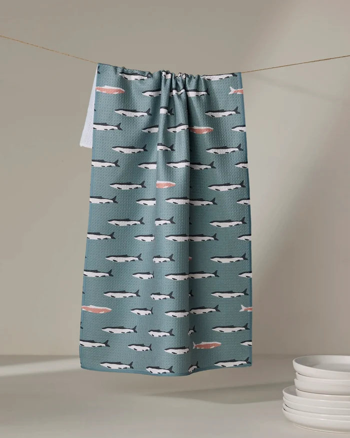 The Lake Fish tea towel by Coit Creative, made from recycled materials, features teal fabric with swimming whales and pink fish. Ultra-absorbent, it's pictured hanging on a line near stacked white plates on a light surface.