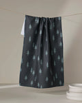 The Coit Creative Lake Pine tea towel, made from recycled materials and featuring a light green pine tree pattern, hangs on a line next to white dishes atop a neutral surface.