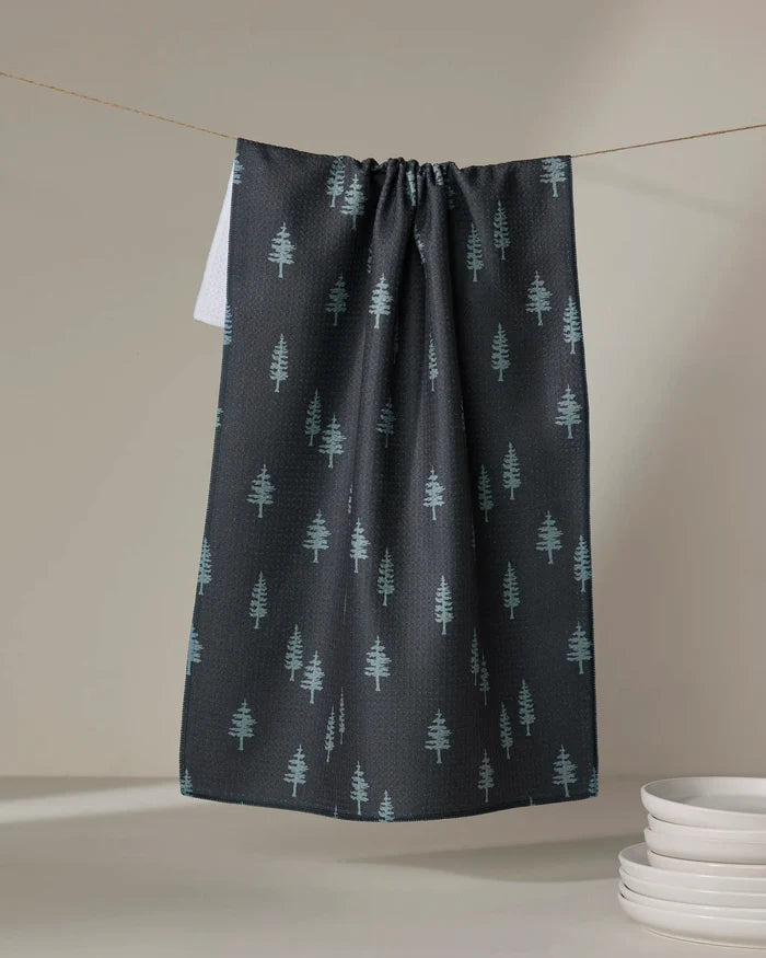 The Coit Creative Lake Pine tea towel, made from recycled materials and featuring a light green pine tree pattern, hangs on a line next to white dishes atop a neutral surface.