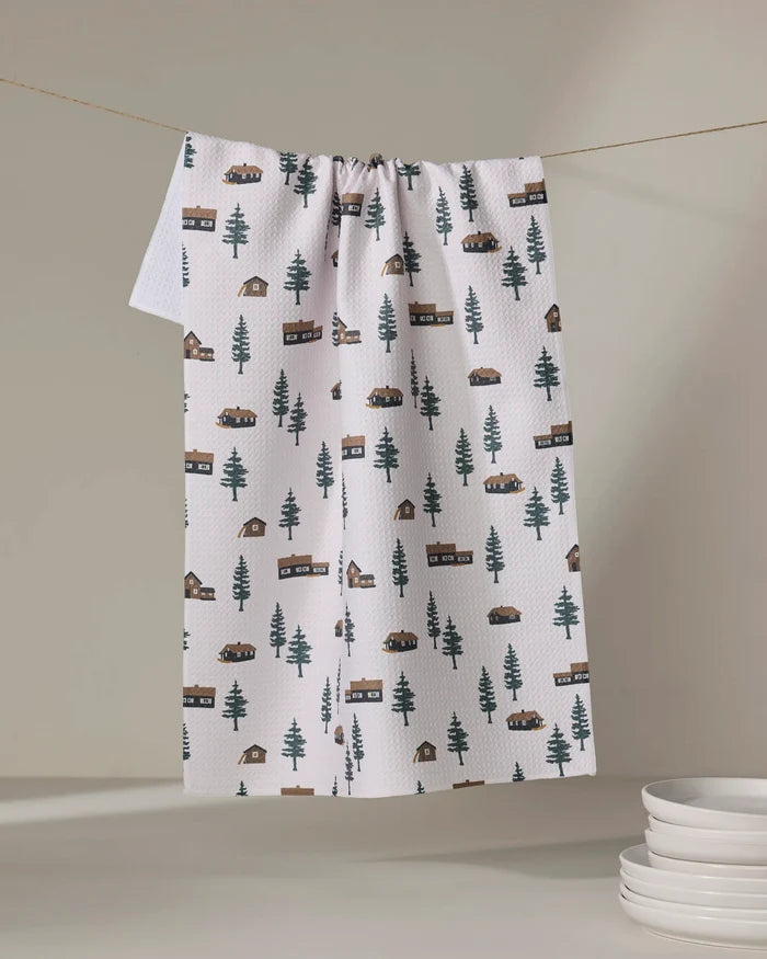 The Coit Creative Lakehouse Cabins tea towel, made from recycled materials, displays a cabins and trees pattern. It hangs on a clothesline above stacked plates, set against a simple neutral background.