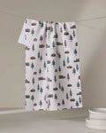 The Coit Creative Lakehouse Cabins tea towel, made from recycled materials, displays a cabins and trees pattern. It hangs on a clothesline above stacked plates, set against a simple neutral background.