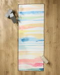 The Sara Mari Lakeside yoga mat, featuring pastel abstract stripes, lies on a wooden floor with a non-slip towel. Two gray blocks sit at the top left and a white water bottle is on the bottom right of the mat.