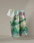 The "Land of Milk and Honey" recycled kitchen towel by Taryn Brennan, featuring an abstract watercolor landscape in green, blue, and pink, hangs on a string against a neutral background.