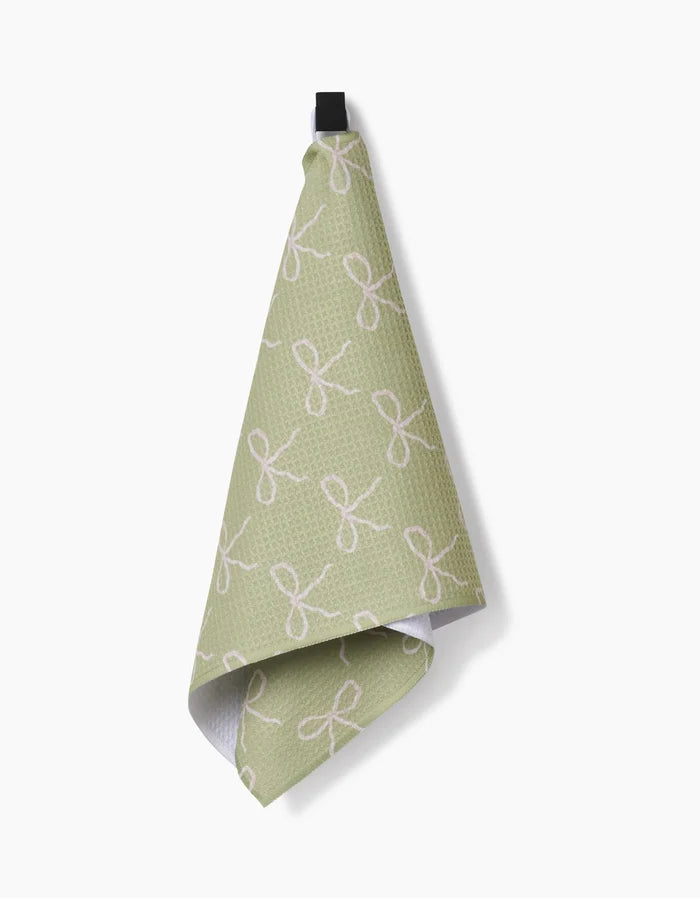 The Laura Prietto Lattice Bows kitchen towel, in light green Geoweave fabric with a white bow pattern, hangs from a black loop against a plain white background.