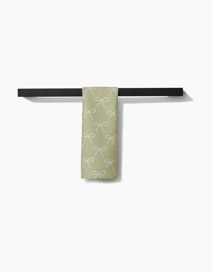The Laura Prietto Lattice Bows Bar Towel, featuring a white bow pattern on light green ultra-absorbent Geoweave fabric, is draped over a modern black towel bar set against a plain white background.