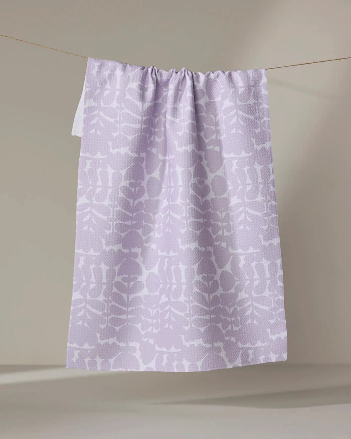 The Katie O'Shea Lavender Scandi Flowers tea towel, featuring a white botanical pattern and made from quick-drying recycled materials, hangs on a thin clothesline against a neutral beige background.